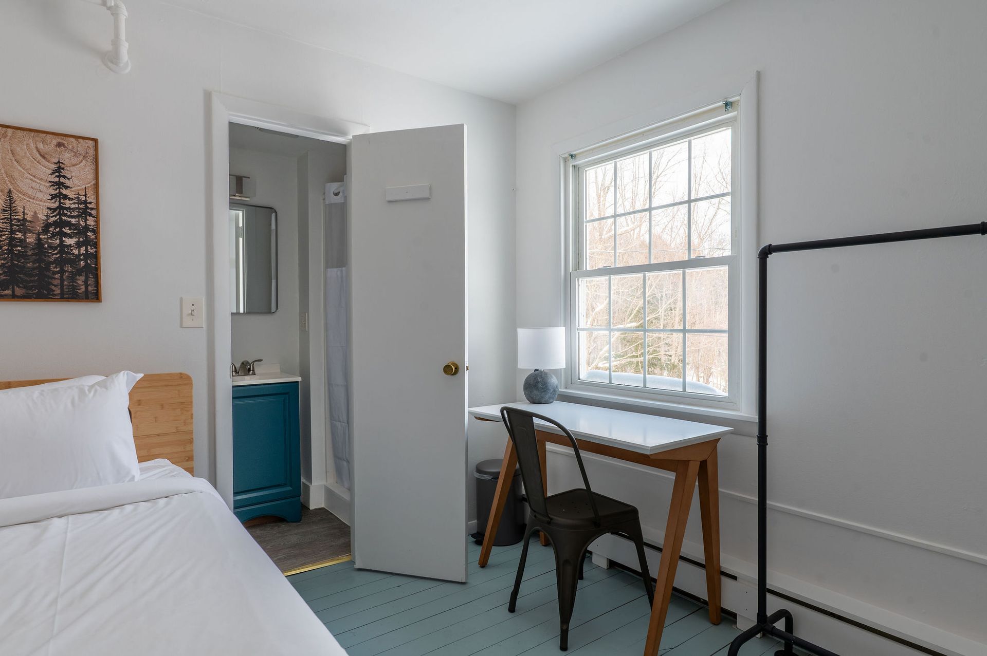 Bedroom with a bed, desk, bathroom doorway, and a window overlooking trees.