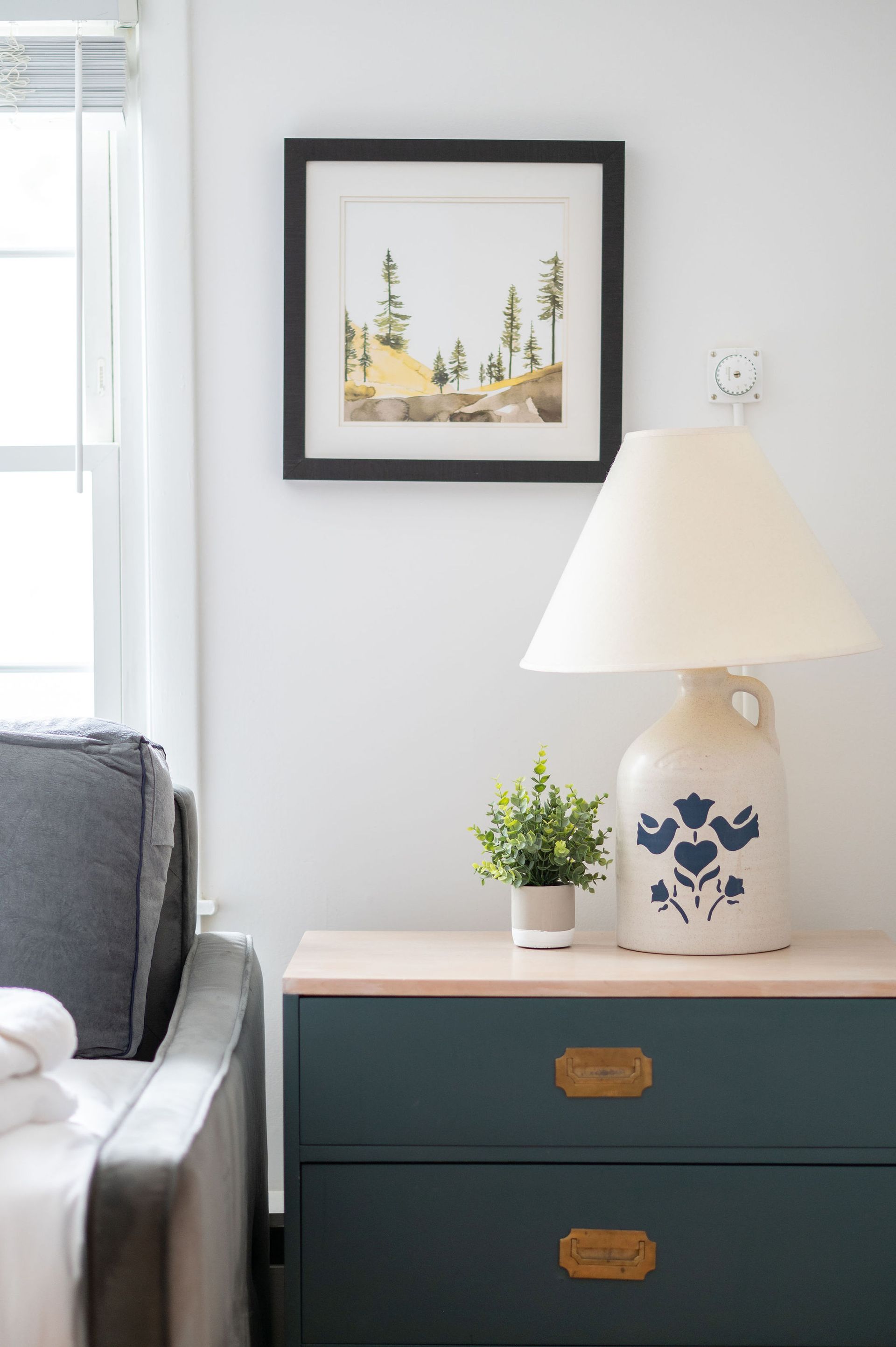 Blue dresser with lamp, framed art, and small plant on top, beside a window and sofa.