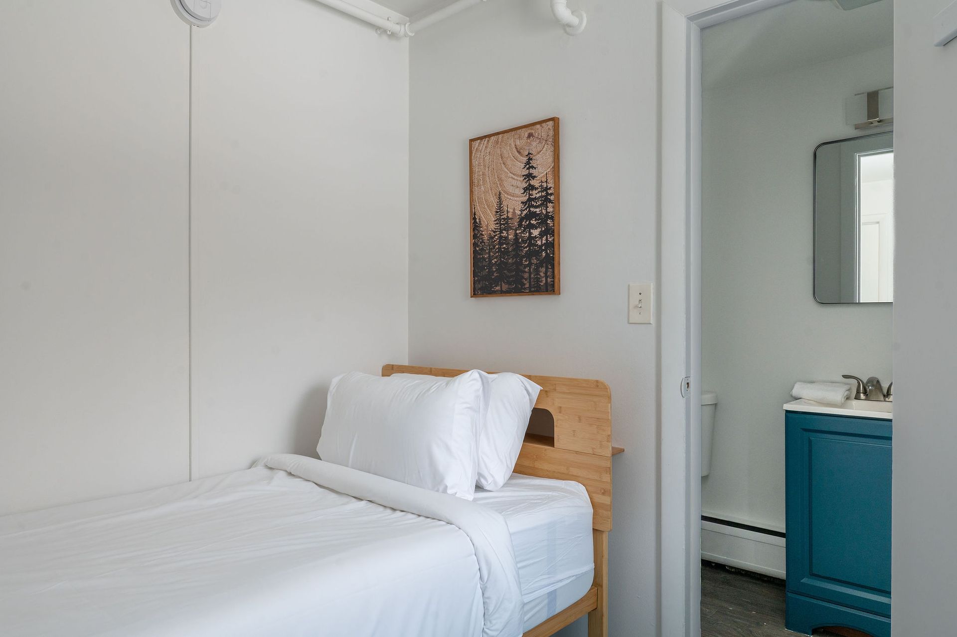 Small bedroom with a single bed, artwork, and an open doorway to a bathroom with a blue vanity.