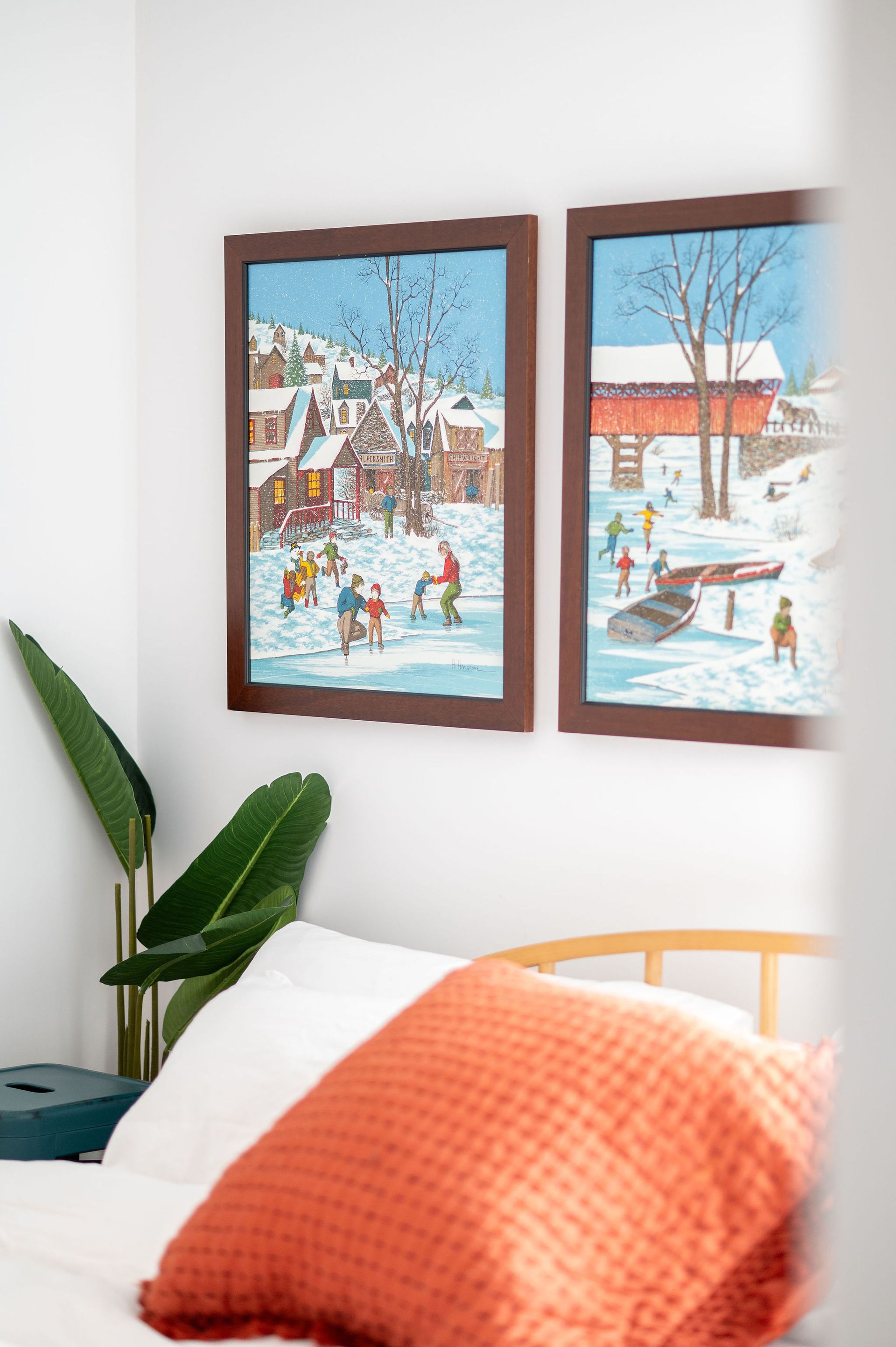 Two framed winter landscape paintings above a bed with orange pillow.