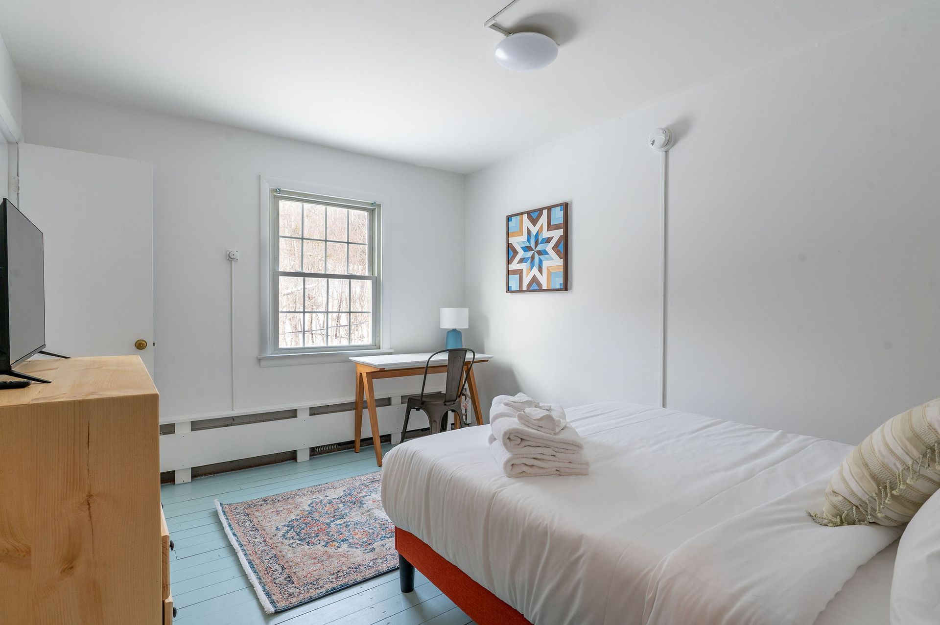 Bedroom with bed, desk by window, rug, and dresser. White walls and blue floor.