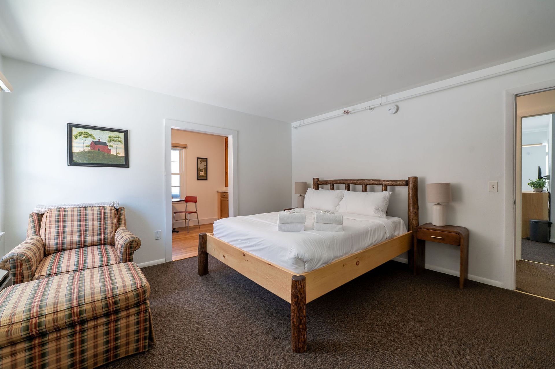 Bedroom with a wooden bed, patterned armchair, and side table. Doorways lead to other rooms.