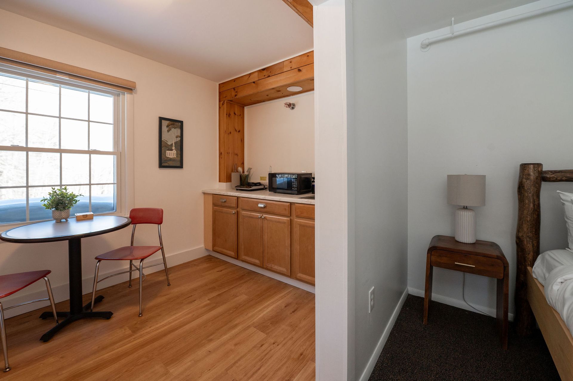 Small room with a kitchenette, dining table, and bed. Wooden cabinets, a window, and a lamp are visible.