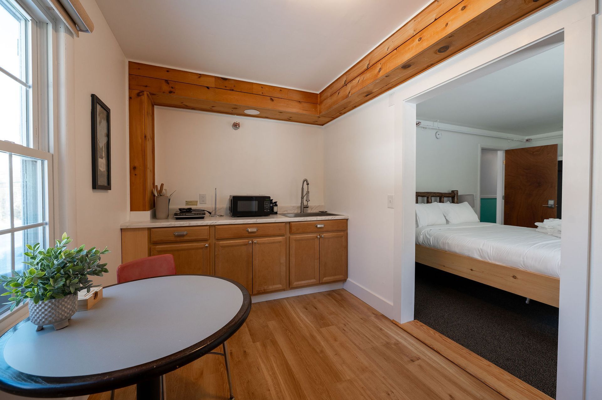 Small kitchen area with table, leading into a bedroom. Kitchen has wood cabinets, microwave and sink. 
