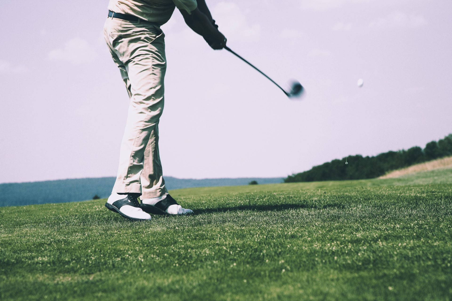 Golfer swinging, hitting a white ball on a green course, blue sky backdrop.