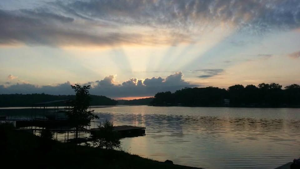 Sunset over a lake; sun rays piercing through clouds, reflecting on water. Dark silhouetted shoreline and docks.