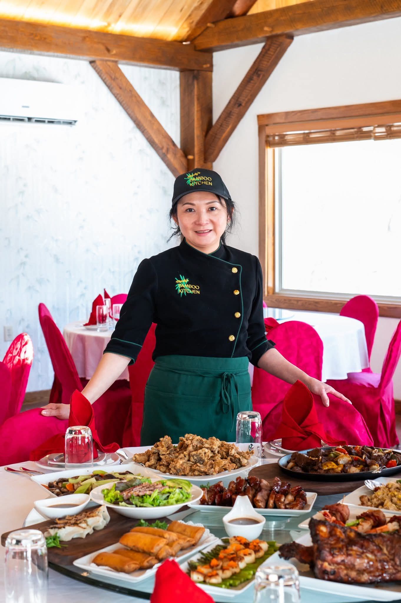 Woman in a chef's uniform standing in front of round table with a white table cloth and red chairs