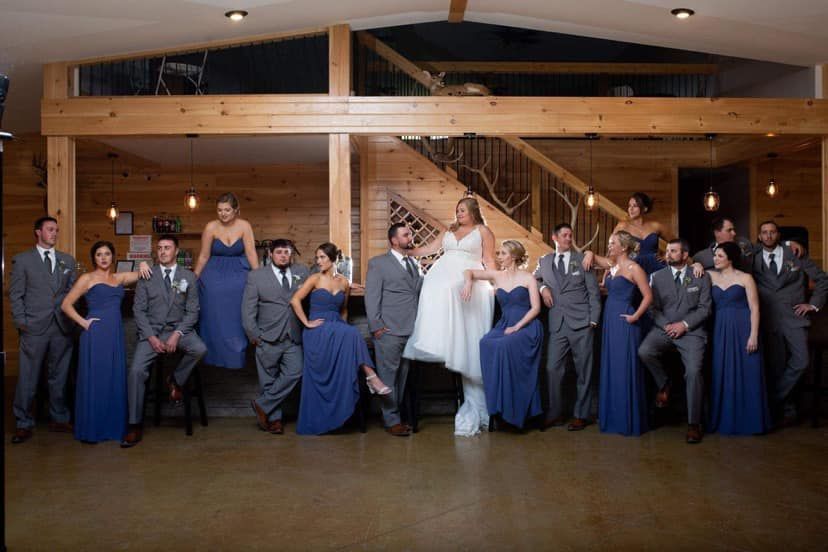 Wedding party: Bride in white dress, bridesmaids in blue gowns, groomsmen in gray suits, posing in a rustic venue.