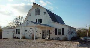 White barn building with a covered entrance, a black roof, and an arched window.