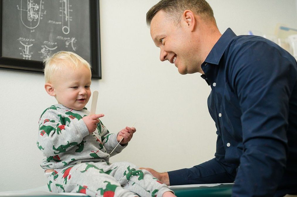 A doctor smiles at a toddler holding a tongue depressor in an examination room.