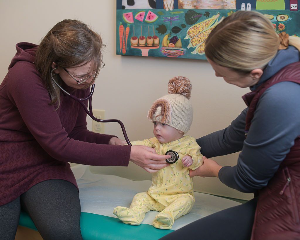 Doctor listens to a baby's chest with a stethoscope while another person supports the baby.