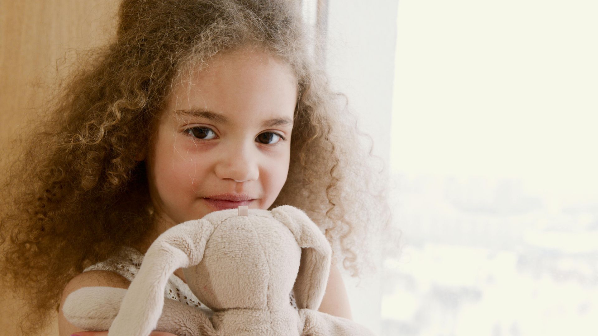 A girl  with curly hair smiles while holding a plush rabbit toy near a bright window.