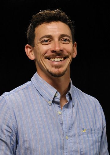 Man in blue t-shirt with logo, smiling, dark background.