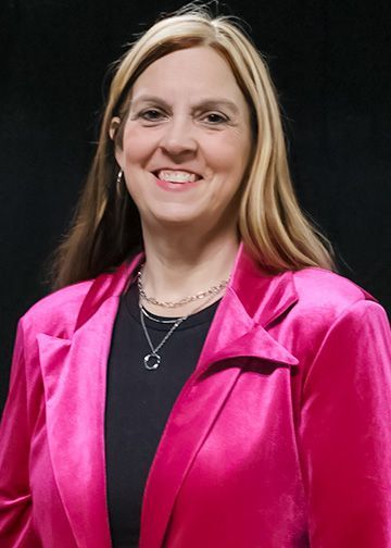 Woman with long brown hair, wearing a black blazer and patterned top, smiling against a black background.