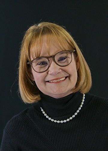 Woman with short brown hair, smiling, wearing a pearl necklace and navy top, black background.