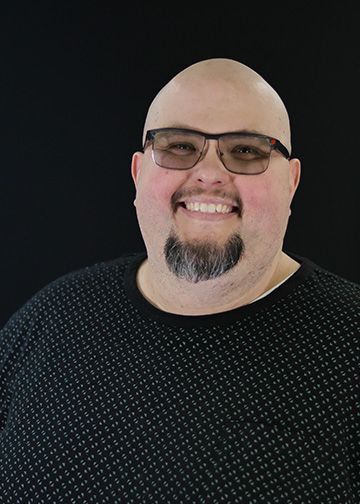 A smiling, bald man with glasses and a goatee stands in front of a black backdrop, wearing a black T-shirt.