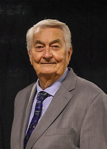 Older man with glasses and white hair, wearing a light blue shirt, smiles. Black background.