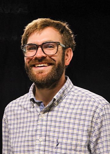Man with beard and glasses wearing a plaid shirt, smiling with hands in pockets, against a dark background.