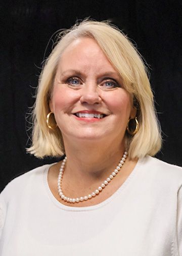 Blonde woman with blue eyes smiles at the camera, wearing a pearl necklace and a light-colored blouse.