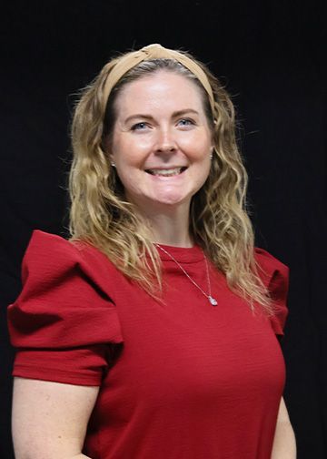 Woman with fair skin smiles at the camera. She wears a black top and a necklace against a dark background.