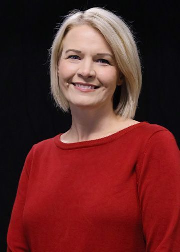 Blonde woman with blue eyes and a subtle smile, wearing a burgundy top against a black backdrop.