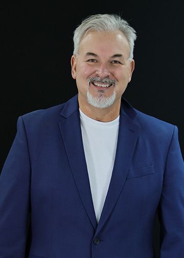 Smiling man with silver hair and beard, wearing a tan jacket and white shirt, set against a black background.