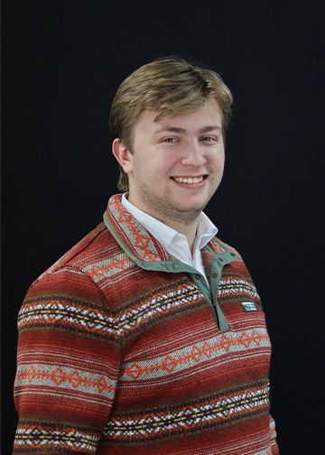 Man with blonde hair, smiling, wearing a brown corduroy blazer over a patterned button-down shirt, in front of a gray wall.