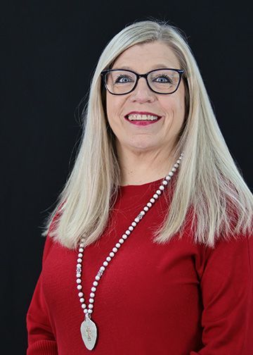 Blonde woman in a light blue top smiles, wearing a necklace, against a black backdrop.