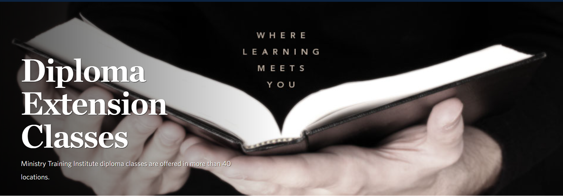A tablet rests on a stack of black books on a wooden desk in a classroom.