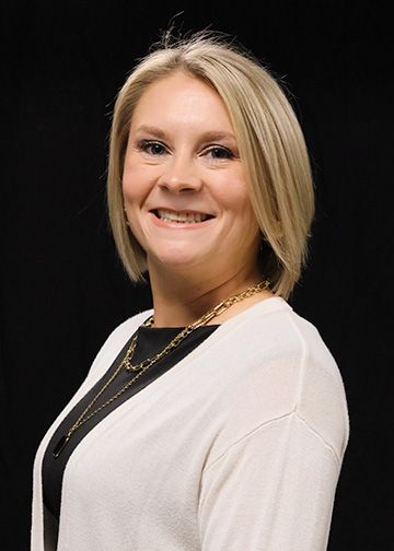 Woman with blonde hair, smiling, wearing a black dress and a gold necklace, against a white background.