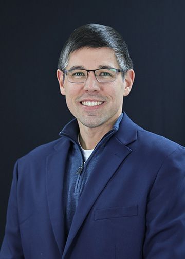 Man with glasses in a blue blazer smiles. Black background.
