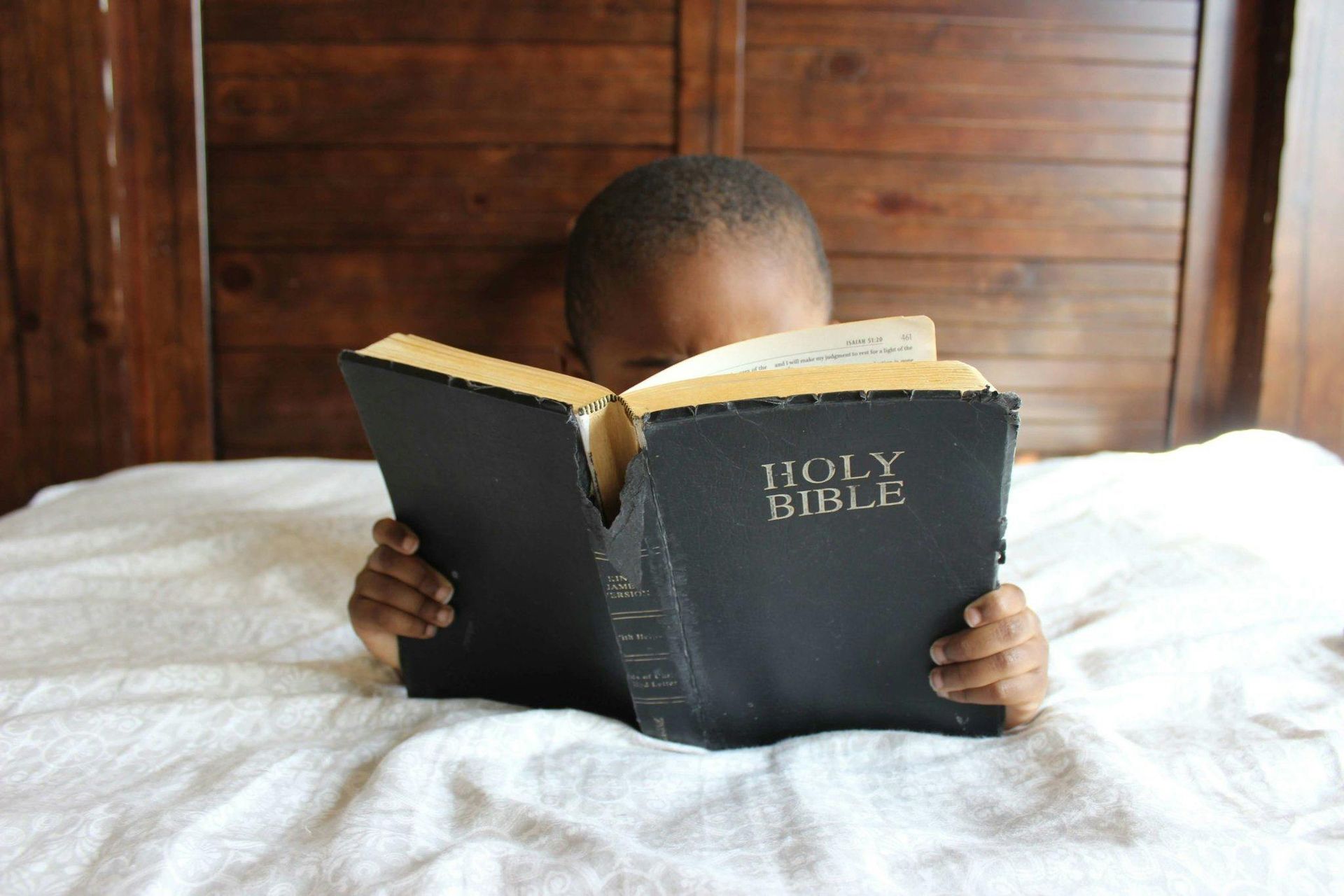 Black child reading the Bible in bed, focused, with wooden headboard behind.