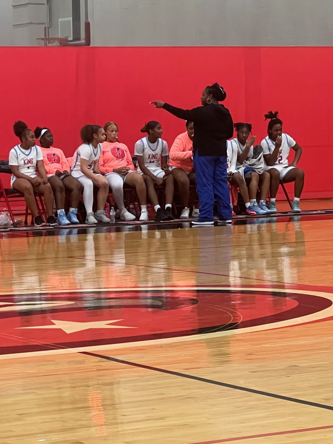 Basketball coach instructing players seated on a bench in a gym. Coach wears blue pants and points to the court.
