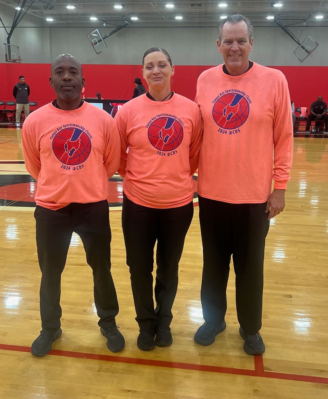 Three people in orange shirts with a logo on a basketball court, standing shoulder-to-shoulder.