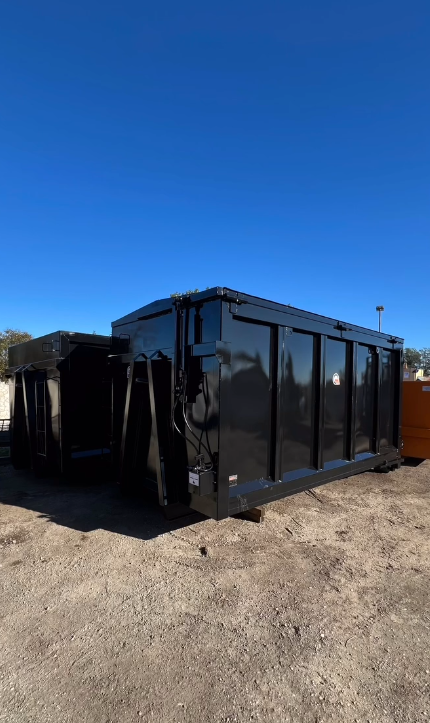 Un grande cassonetto nero è appoggiato in cima a un campo sterrato.