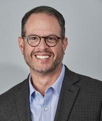 Man with glasses smiles, wearing a blue shirt and checkered blazer against a gray background.