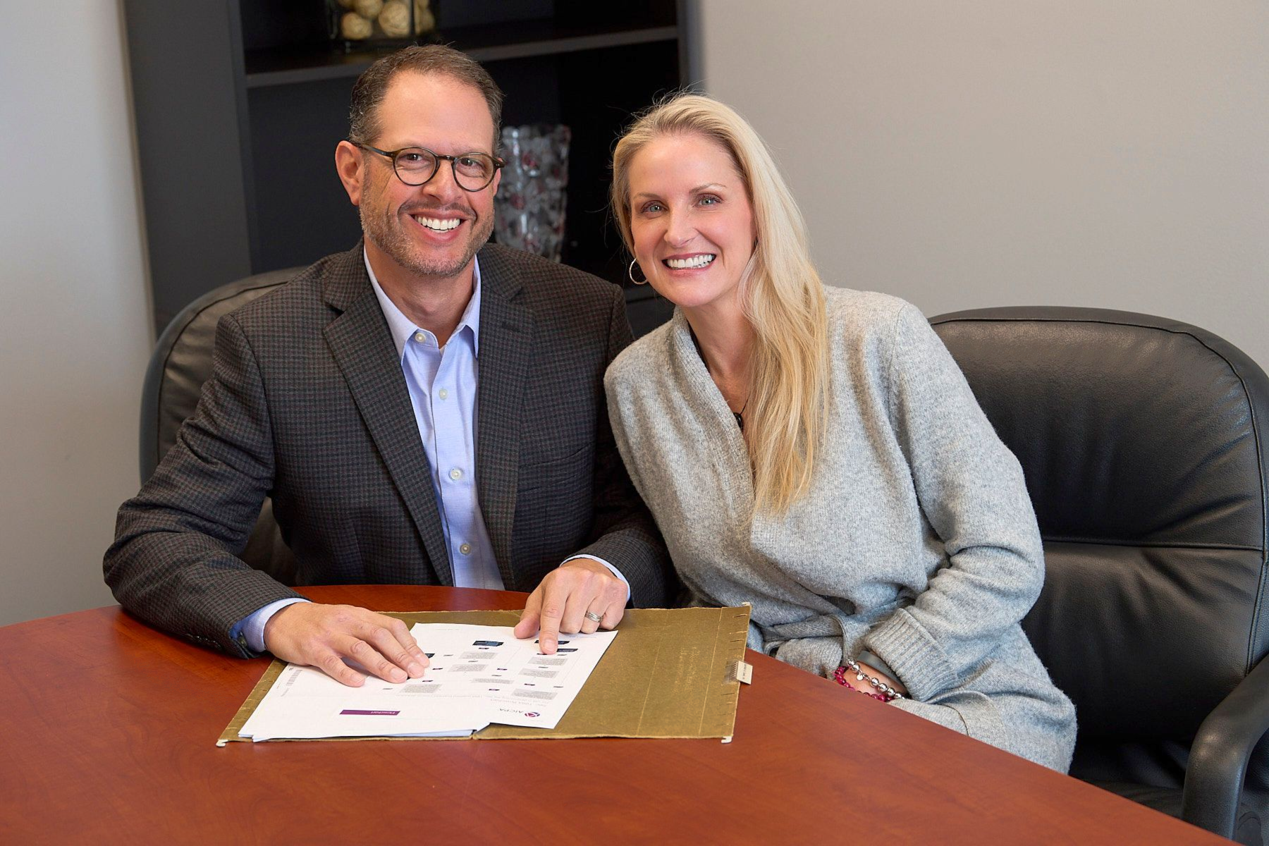 Man and woman smiling, sitting at a table with papers, possibly a business setting.