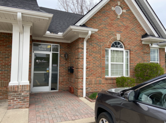 Exterior of a brick building with a glass door, address visible. A car is parked to the right.