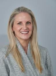 Woman with long blonde hair, smiling, wearing a gray sweater, standing against a gray background.
