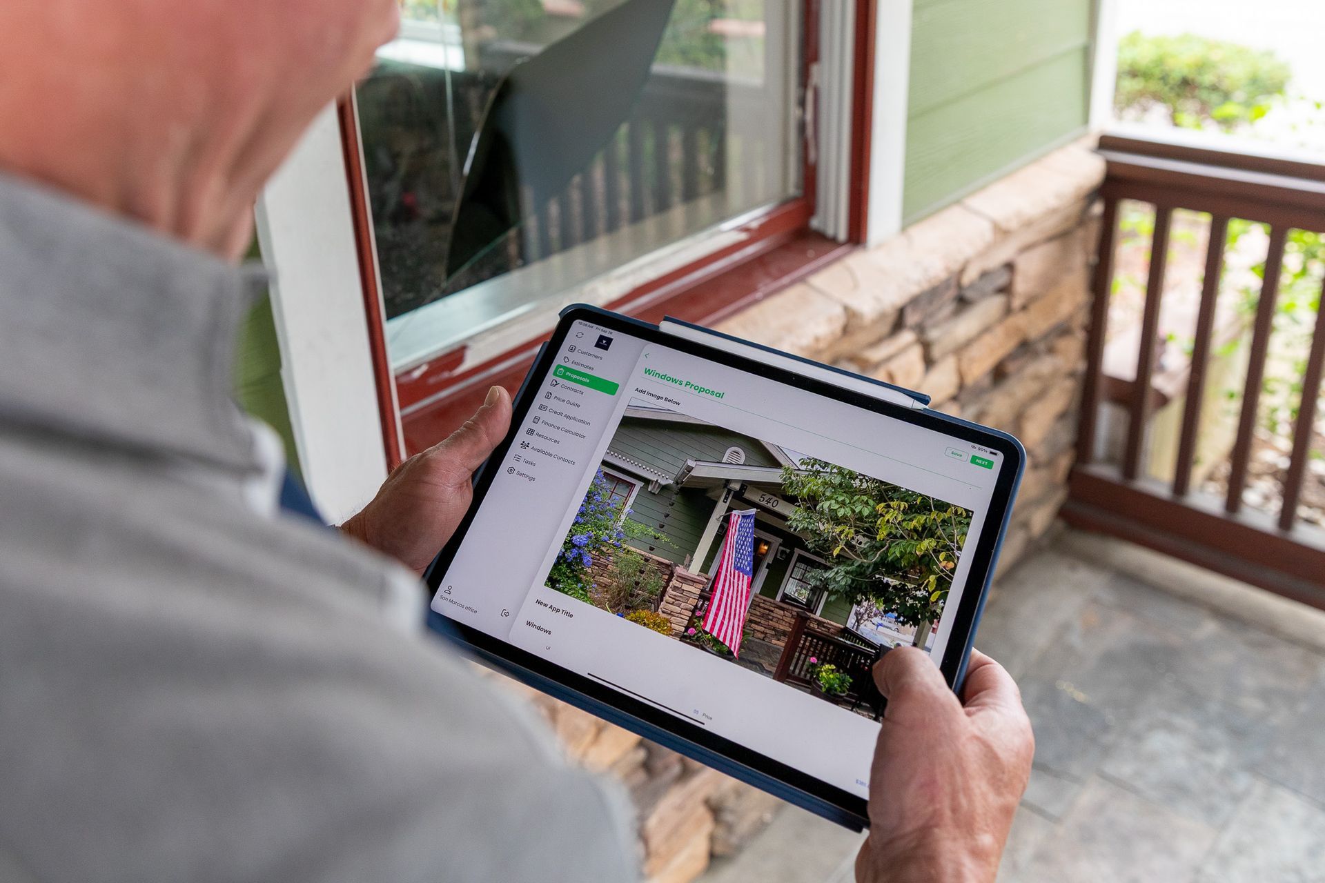 Person holding a tablet, looking at a photo of a house, standing near a porch with brick and wooden features.