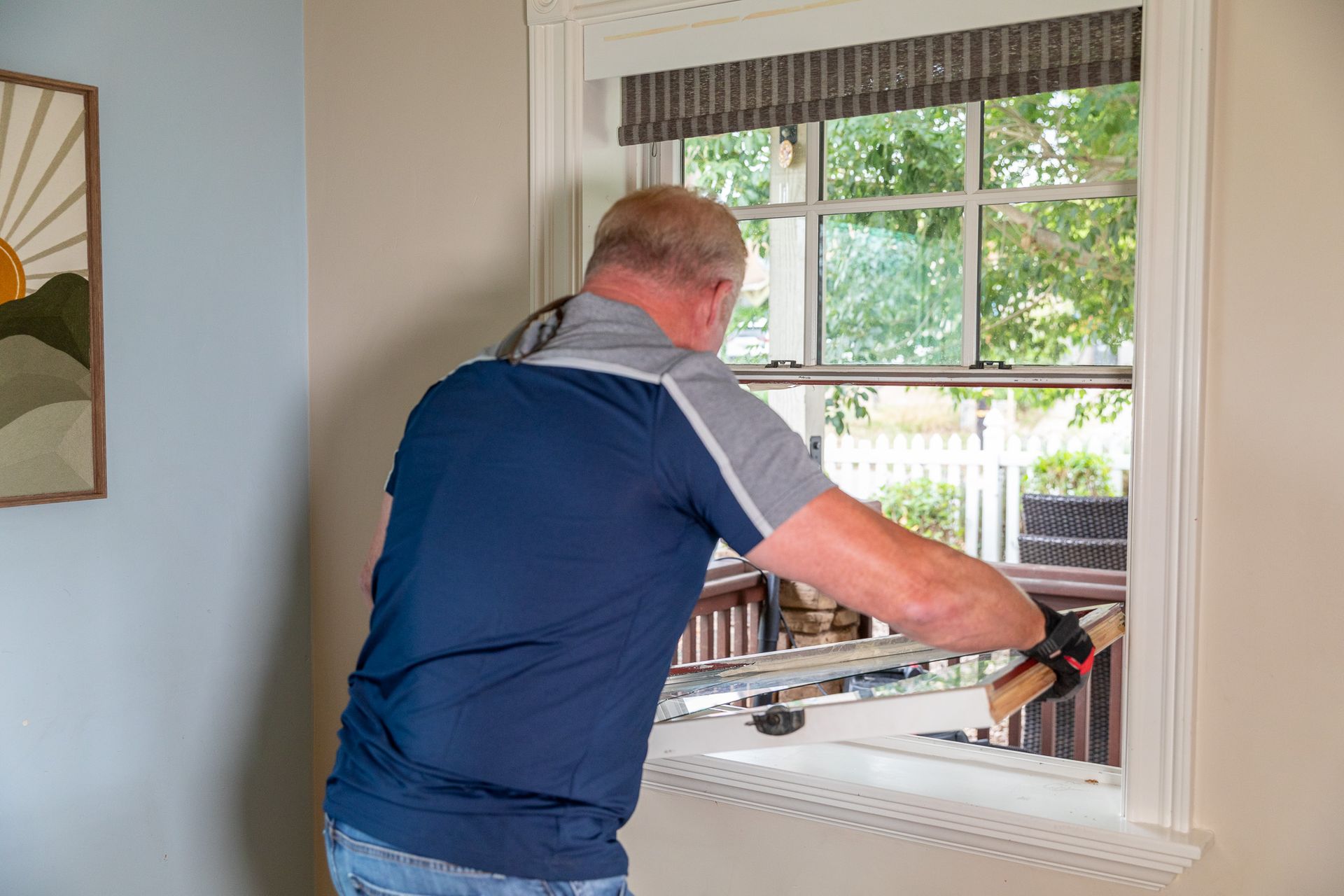 Man opening a double-hung window. He wears a gray and blue shirt and is in a room with a framed art.