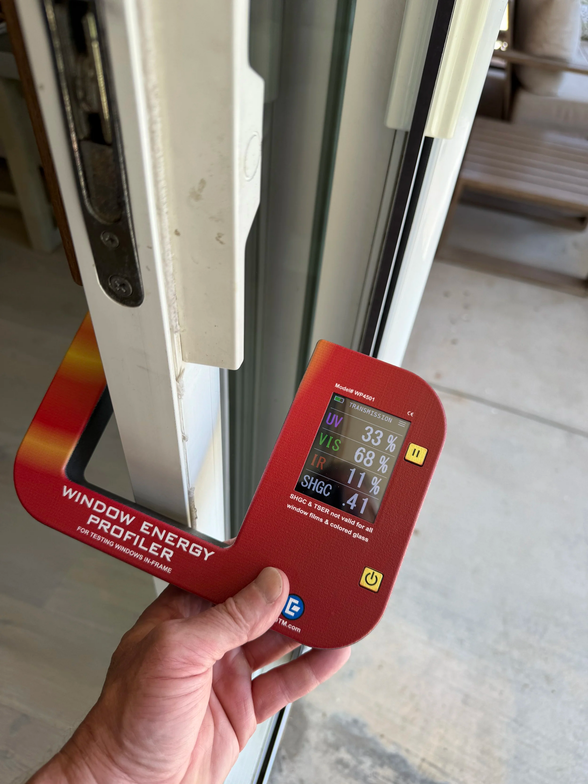 A hand holds a red window energy profiler against a white window frame, displaying energy efficiency data.