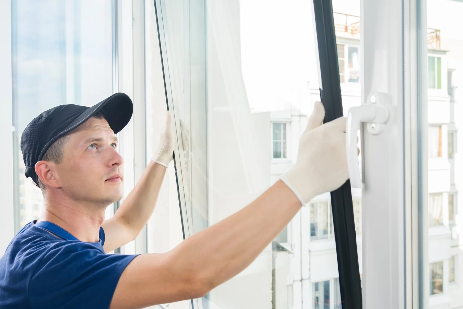 Man in cap and gloves installing a window, looking upwards. White frame, daylight setting.