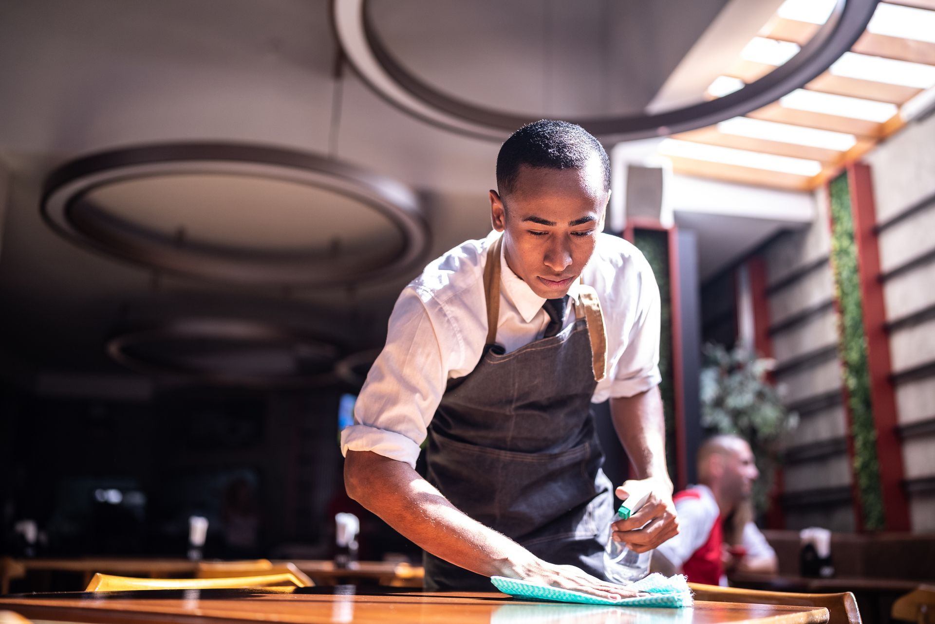 A man in an apron is cleaning a table in a restaurant.