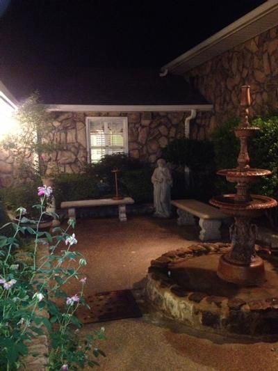 Stone patio with fountain, benches, statue, and flowering plants; lit at night.