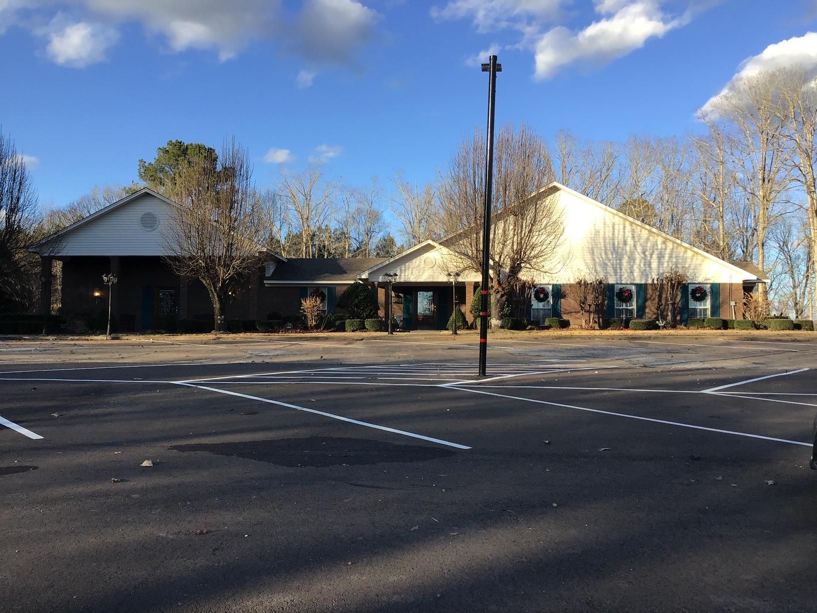 Parking lot in front of a low brick building with two peaked roofs and several windows, sunny day.