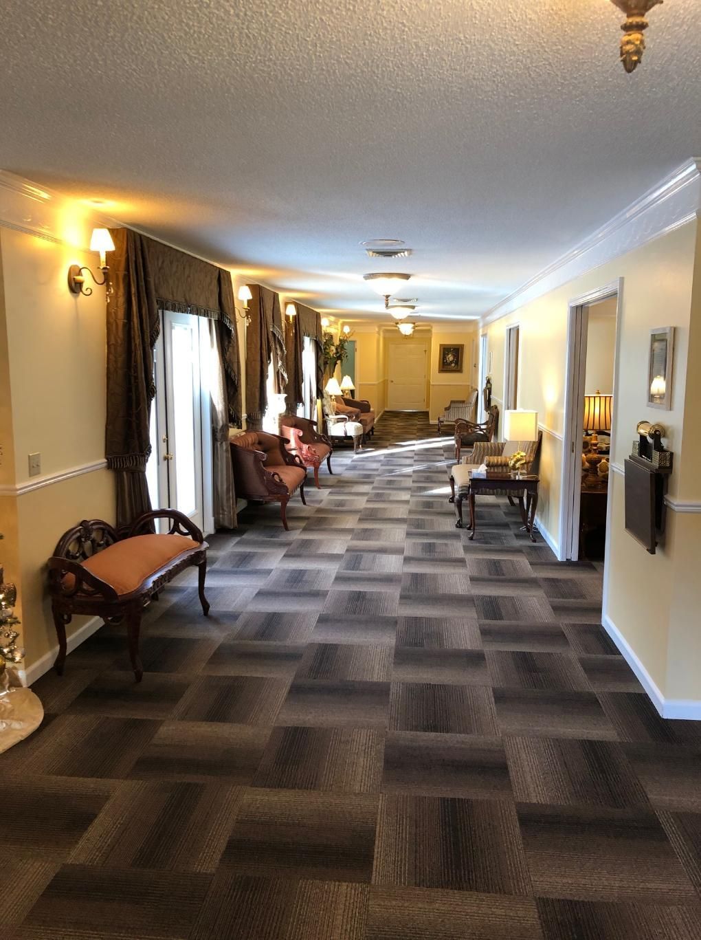 Long hallway with patterned carpet, antique furniture, and neutral-toned walls, at a memorial service or funeral home.
