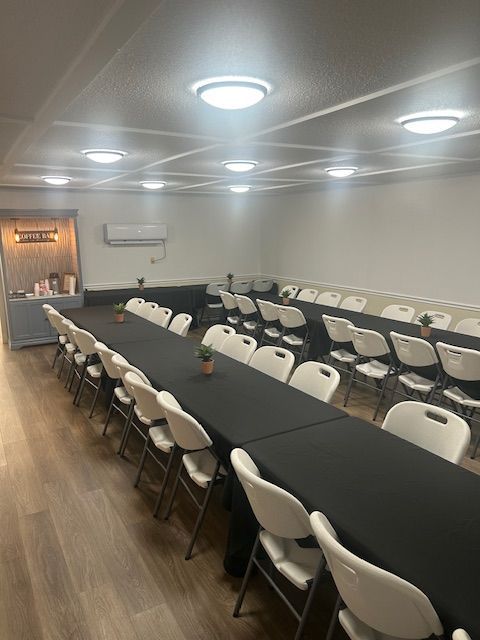 Indoor event space with long tables covered in black linens, white chairs, and overhead lighting.