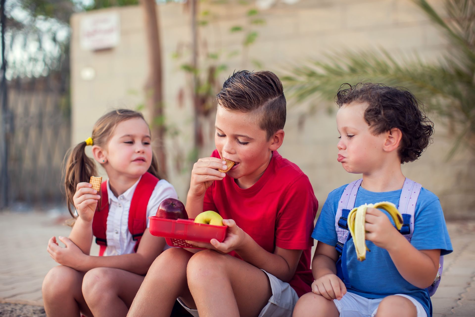 Three children are sitting on the ground eating fruit.