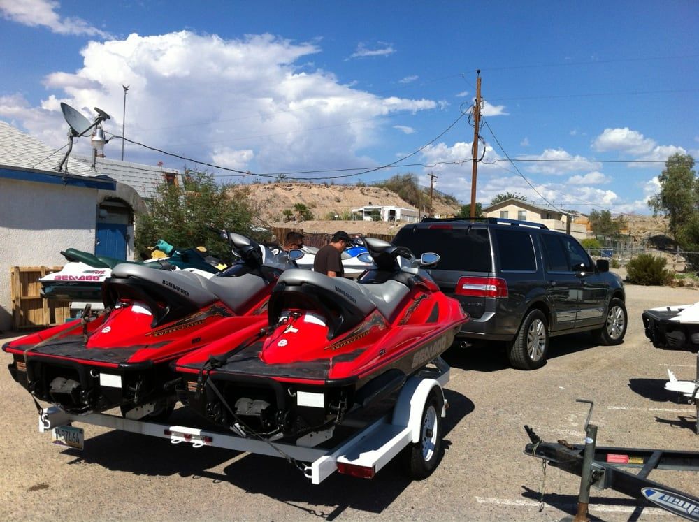 Two red jet skis on a trailer beside a black SUV in a dusty lot under a blue sky